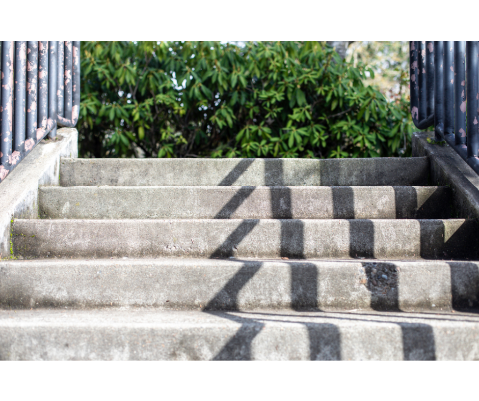 Concrete steps with black stripes leading up to a metal railing with greenery in the background.